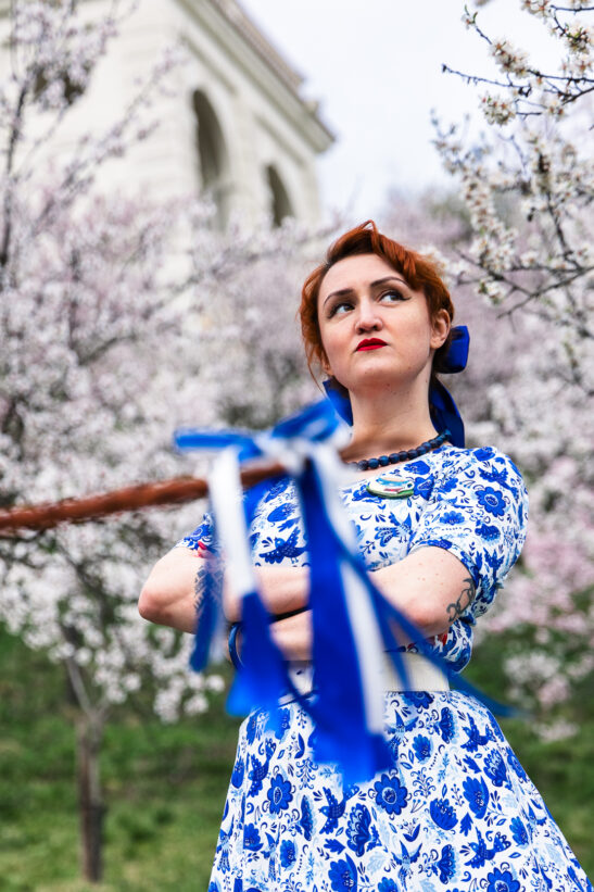 Woman with red hair in a blue floral dress stands with arms crossed, blue ribbons blowing in front, blooming trees in the background.