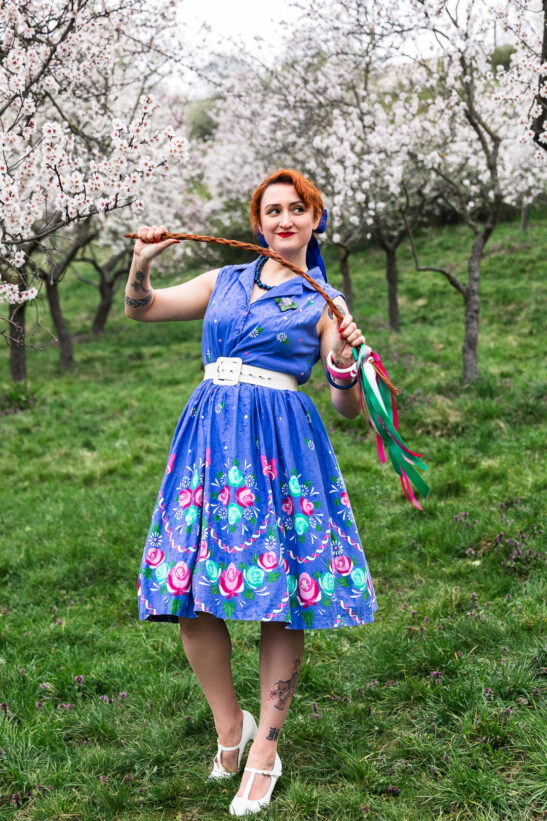 Woman in a blue floral dress with a white belt stands in a blossoming orchard, holding a braided rope with ribbons.