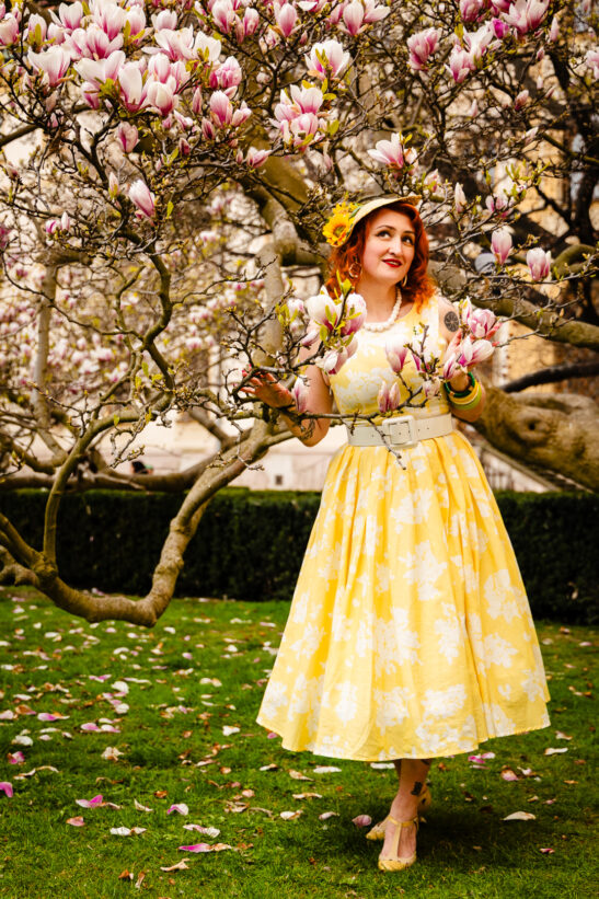 Woman in a yellow floral dress and hat stands among pink magnolia blossoms in a garden.