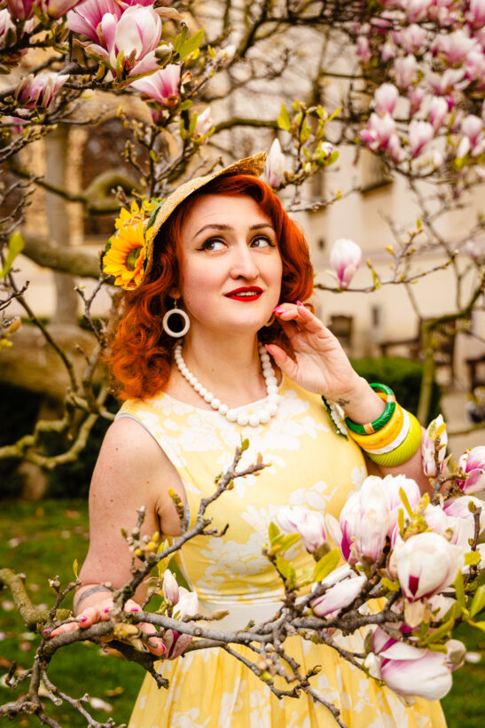 Woman with red hair in a yellow floral dress stands among magnolia blossoms, wearing a straw hat with a sunflower and a white bead necklace.”,