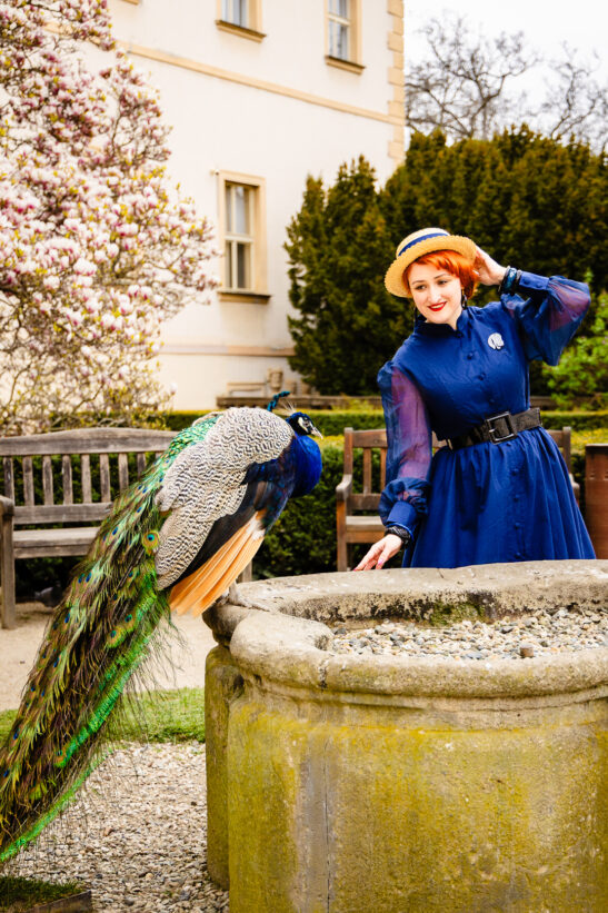 Woman in a blue dress and straw hat standing beside a peacock perched on a stone well in a garden setting.