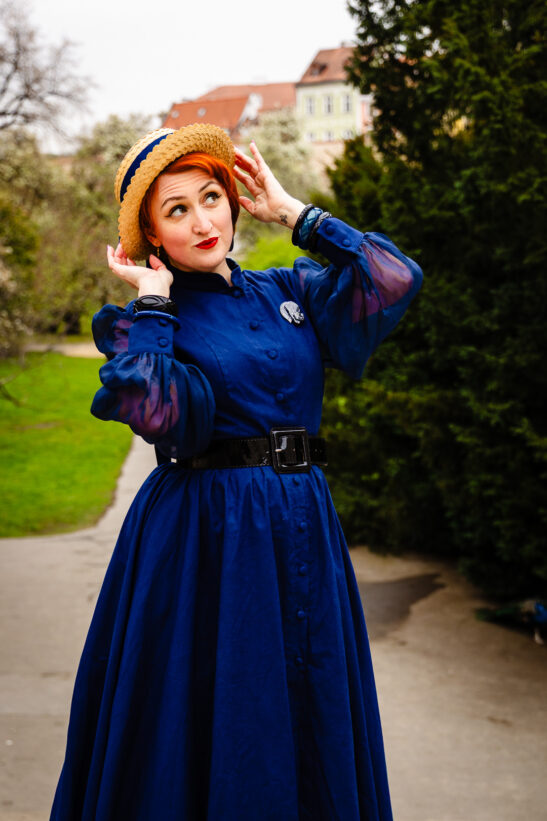 Woman in a royal blue vintage dress with a straw hat, posing outdoors on a park path.