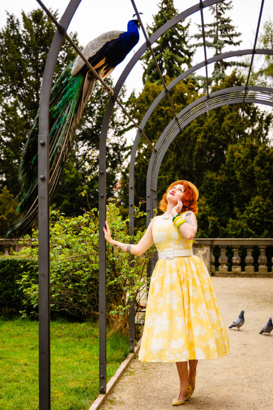 Woman in a yellow floral dress and heels stands in a garden, looking up at a peacock perched on a curved metal arch above her.