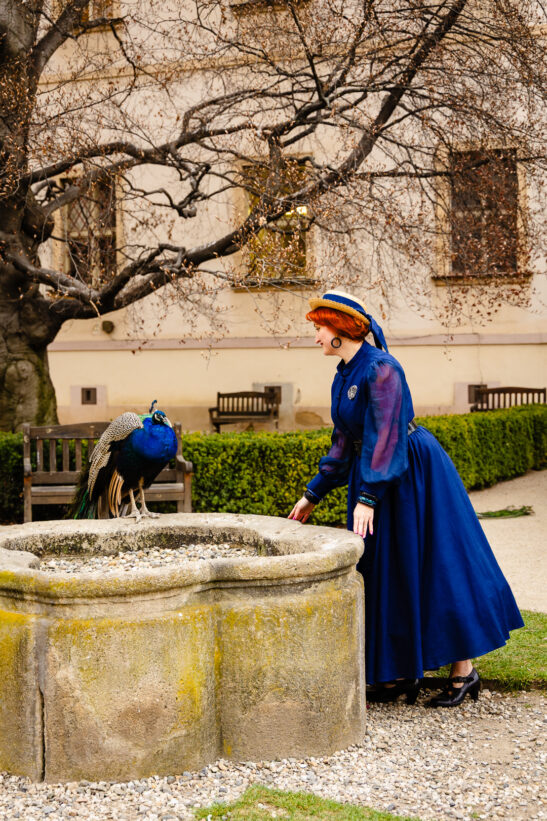 Woman in a cobalt blue vintage dress and straw hat bending toward a stone well as a peacock perches on the edge in a garden courtyard.
