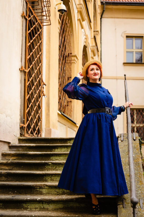 Woman in a vintage blue dress and straw hat posing on stone steps outside a yellow building with iron window bars, looking to the side.