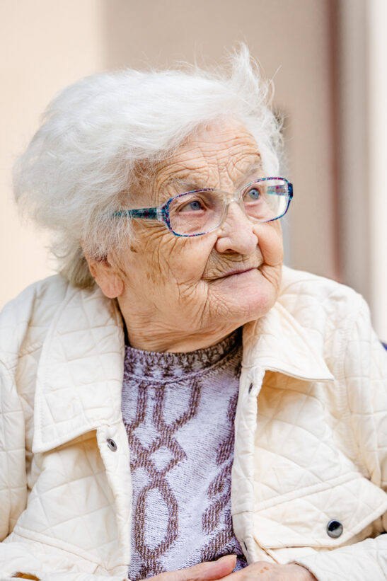 Elderly woman with white hair and colorful glasses looking to the side, wearing a quilted cream jacket and patterned sweater.