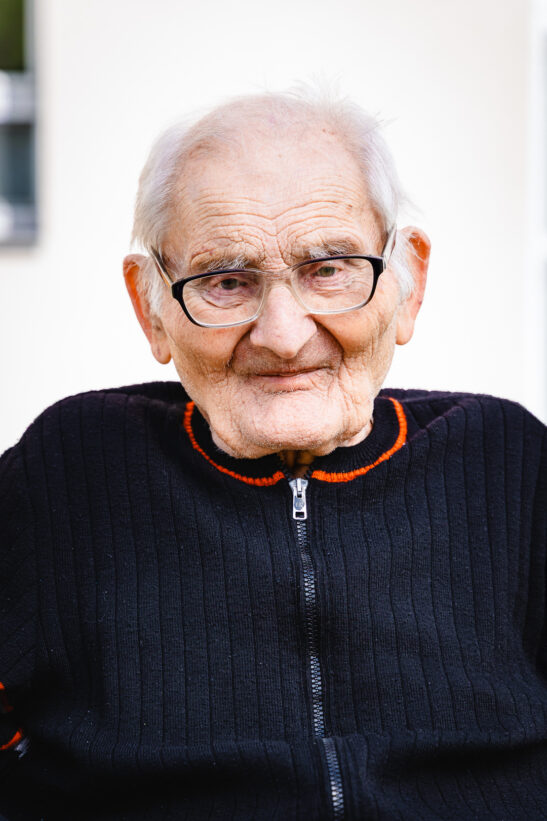Smiling elderly man with white hair and glasses, wearing a dark zip-up sweater with orange trim.