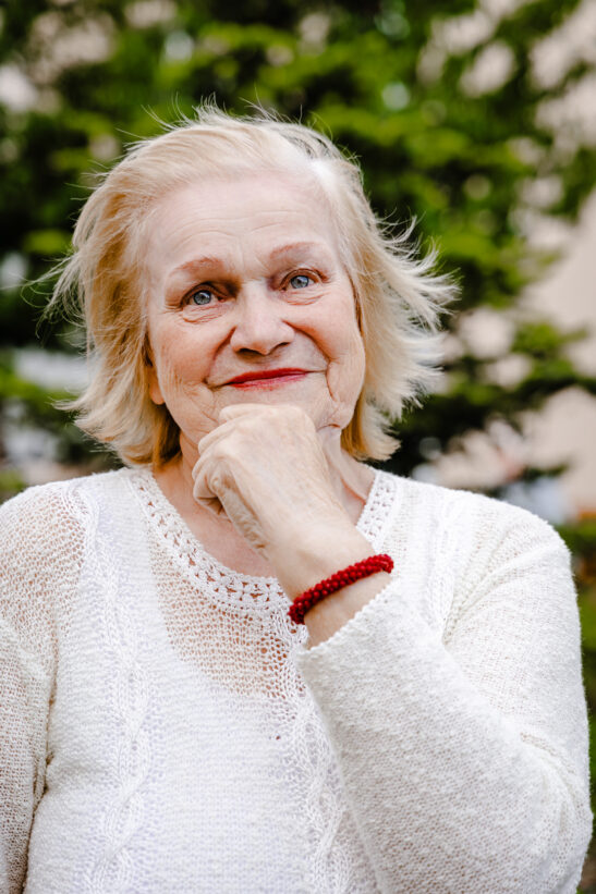 Smiling elderly woman with short white hair in a white knit sweater outdoors, wearing a red beaded bracelet.