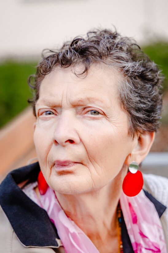 Close-up portrait of an older woman with short curly gray hair, wearing bold red earrings and a pink scarf outdoors.