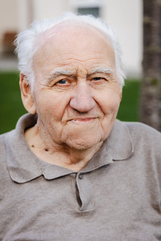 Close-up portrait of an elderly man with white hair, wearing a beige polo, outdoors.