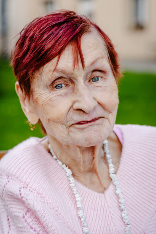 Close-up portrait of an elderly woman with short red hair, wearing a pink sweater and a white beaded necklace, outdoors with greenery behind her.