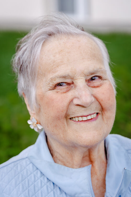 Smiling elderly woman with short gray hair wearing a light blue quilted jacket outdoors.