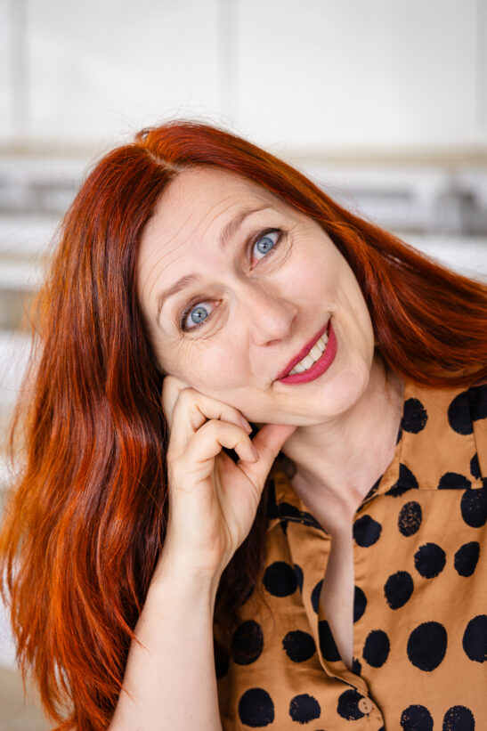 Portrait of a smiling woman with long red hair resting her chin on her hand, wearing a tan blouse with black polka dots.