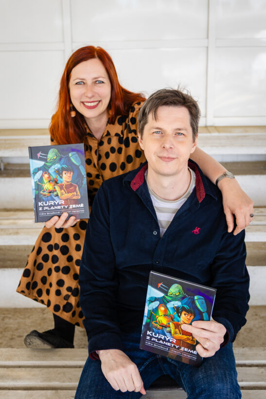 Smiling red-haired woman and man pose on stairs holding copies of a colorful sci‑fi book with illustrated robots and astronauts.