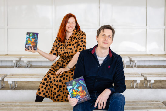Two adults sit on white bleachers; the woman with red hair in a brown polka‑dot dress holds a colorful illustrated book toward the camera, smiling.