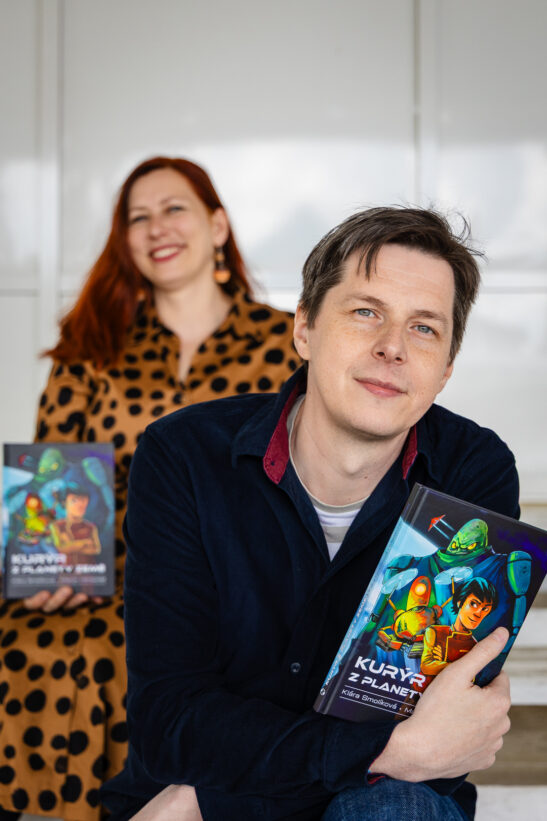 Man in a dark blue shirt holds a colorful illustrated book, with a smiling woman in a leopard‑print dress standing behind him.