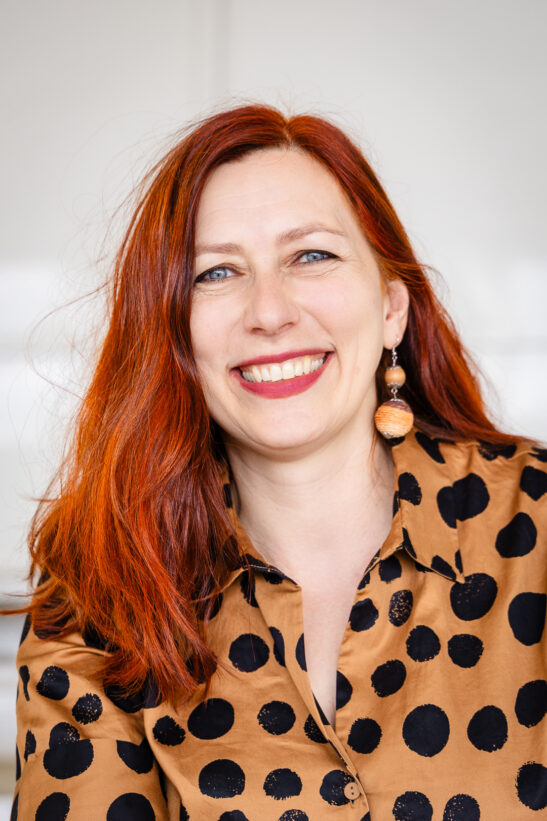 Smiling woman with red hair wearing a tan polka-dot blouse and round earrings, against a light background.